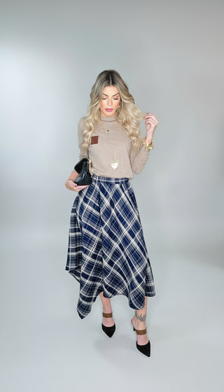 Woman wearing a beige sweater and blue plaid skirt with black shoes indoors.