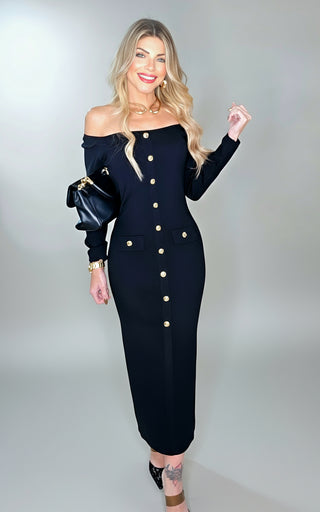 Woman wearing a black off-shoulder dress with gold buttons in an indoor setting.